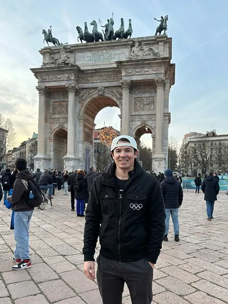 Austin Taylor, wearing a black jacket with Olympic Rings on it, stands in front of the Arco della Pace in Milan, Italy.
