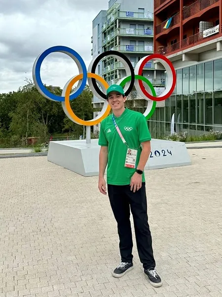 Austin Taylor stands in front of a statue of the Olympic Rings in Paris, France in the Olympic Village. He wears a green shirt and hat with his credentials across his chest.