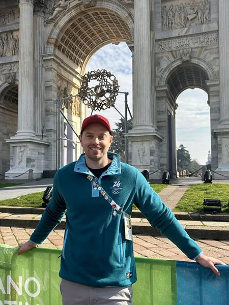 Ben Cooke stands in front of the Arco della Pace in Milan, Italy. He wears his Olympic credentials across his chest and a teal pullover with the 2026 Winter Olympics logo on the chest.