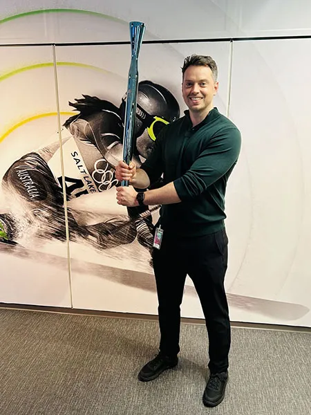 Ben Cooke stands in front of a wall with a skier surrounded by the green rings of Deloitte. He is holding a blue chrome Olympic Torch.
