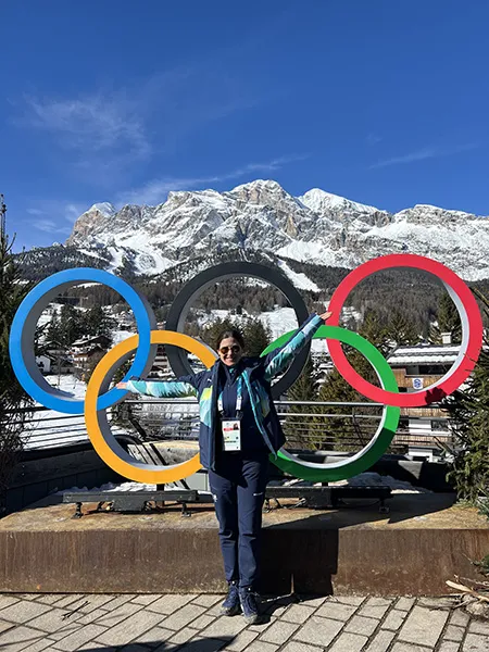 Katrina Galas stands in front of a statue of the multicolored Olympic Rings with a mountain in the background.