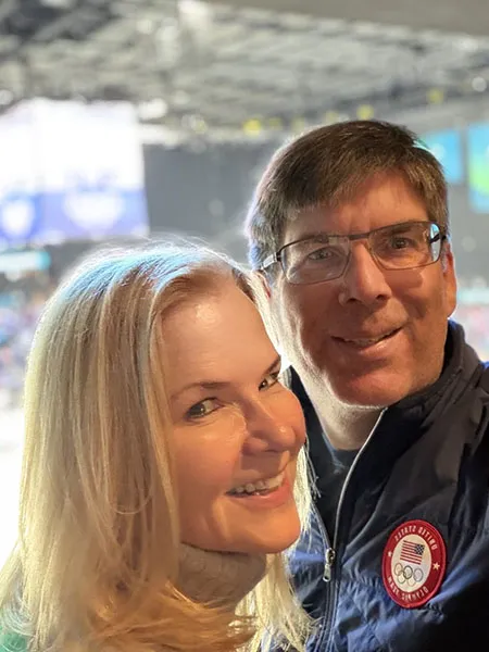 Maureen Sweeney smiles in a selfie with her husband who is wearing a Team USA jacket. They are in an Olympic stadium.