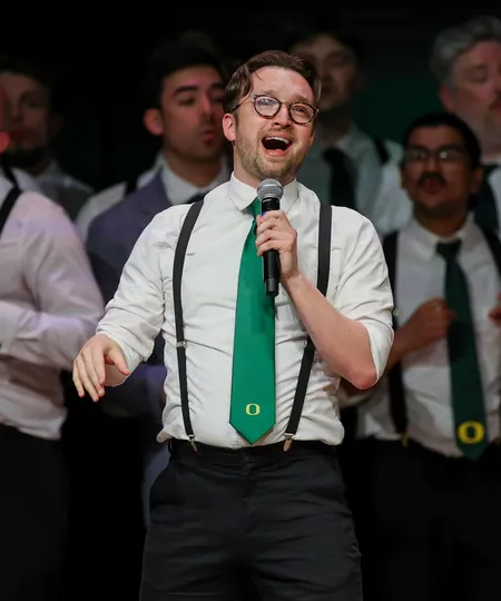 Teigh Bowen wears a white button down shirt, green suspenders and a green tie with an O at the bottom while singing into a microphone at the 25th Anniversary Show for On the Rocks.