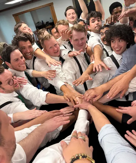 Young and middle aged men in white button down shirts and suspenders huddle together putting their hands together in the middle of the photo.