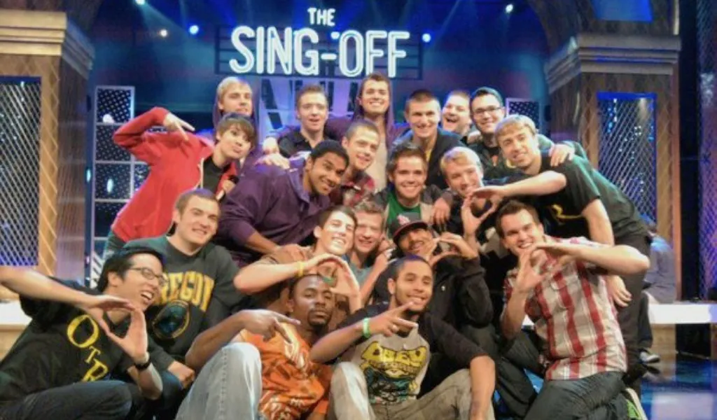 Members of On the Rocks pose with singers from other a cappella groups on stage in front of a bright sign that says The Sing-Off.