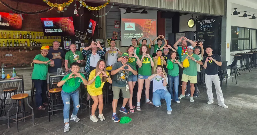 A group of people in Oregon Ducks merch make O shapes with their hands in front of a bar.