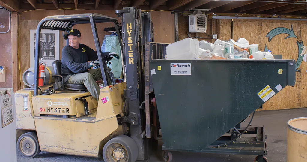 A man drives a forklift carrying a dumpster filled with plastic gallon bottles.