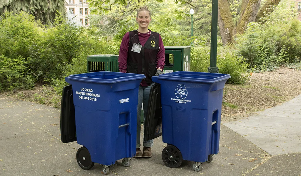 A young woman smiles while holding two blue recycling bins that say UO Zero Waste Program on the sides.