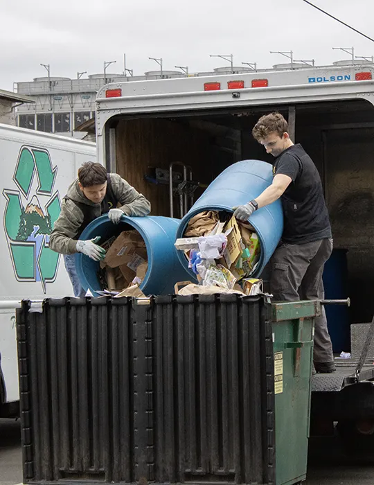 Two young men pour paper out of barrels in the back of a truck into a dumpster below them.