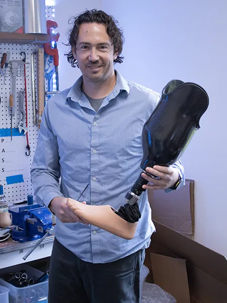 A man holds a prosthetic leg in front of a workshop tool bench.