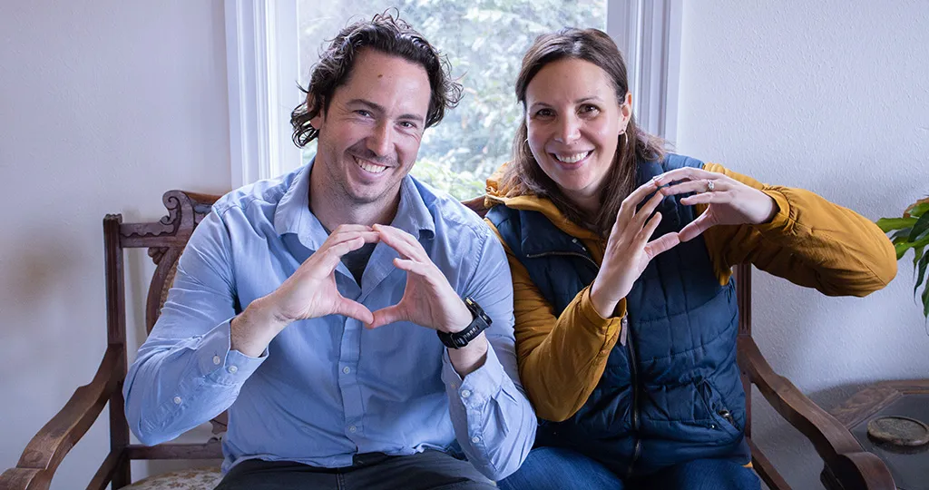 A man and a woman make O shapes with their hands while sitting on a bench in front of a window.