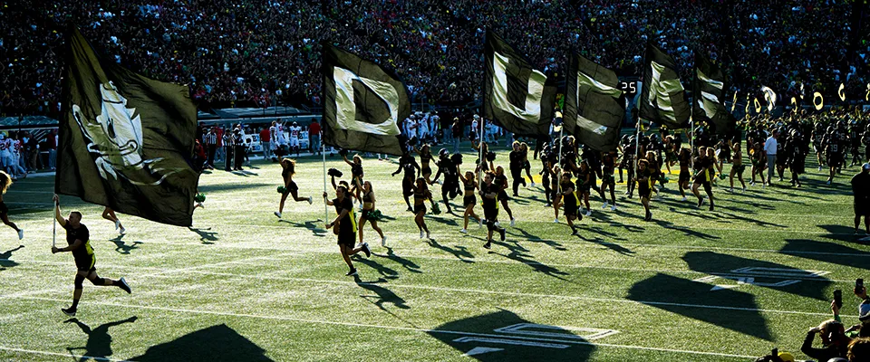 People holding flags that spell out DUCKS run across a football field.