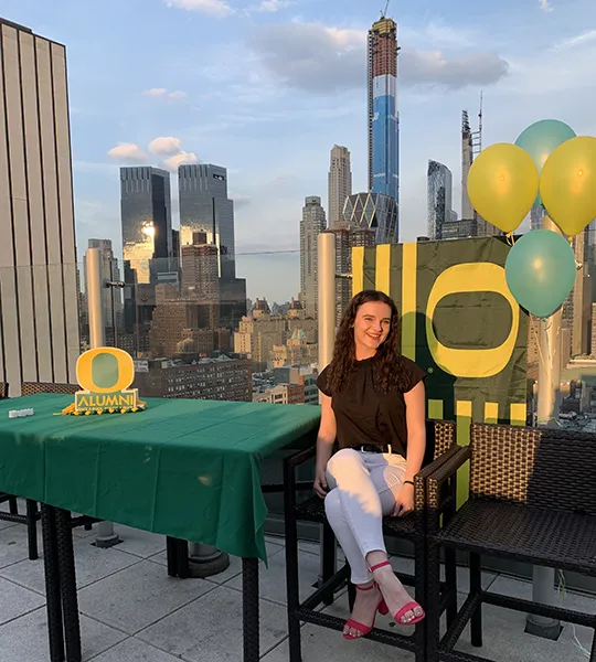 Bailey Holloway sits in a chair next to University of Oregon decorations with the New York City skyline behind her.