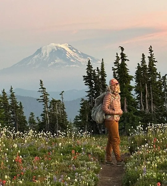 A woman in hiking gear stands in a wildflower field in front of green trees and a snow-capped mountain in the distance under a cloudy pink sky.