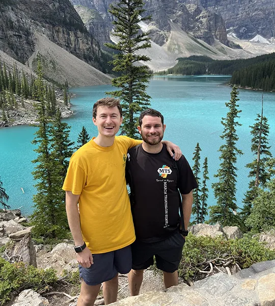 Two men stand smiling in front of a bright blue lake surrounded by gray mountains and green trees.