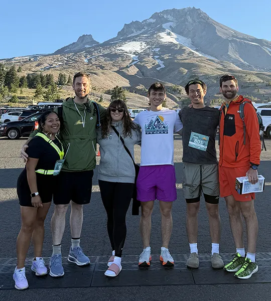 A group of six people stand with their arms around each other in front of a mountain in running gear.