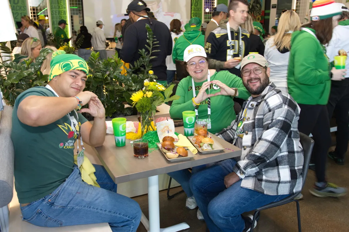 Three people in Oregon Ducks gear sit at a table with food on it and smile at the camera.