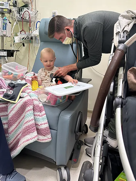 A man holds a stethoscope to a toddler's chest in a hospital room.