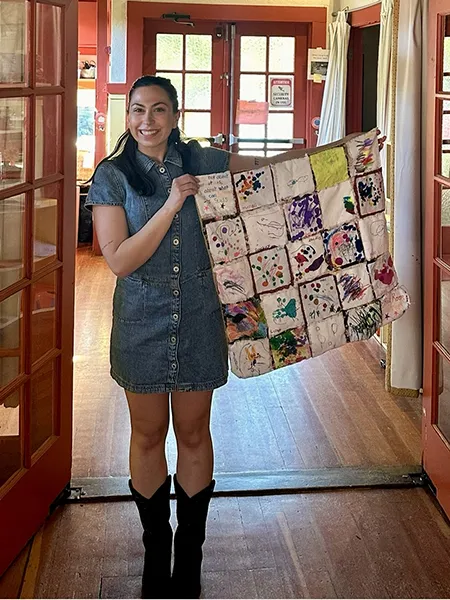 A woman in a jean dress holds up a small quilt with finger paintings on each square.