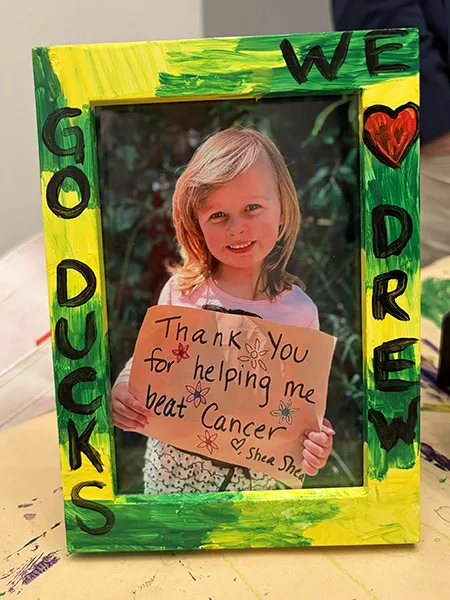 A hand-painted green and yellow picture frame around a photo of Shea holding a sign that says Thank you for helping me beat cancer. 