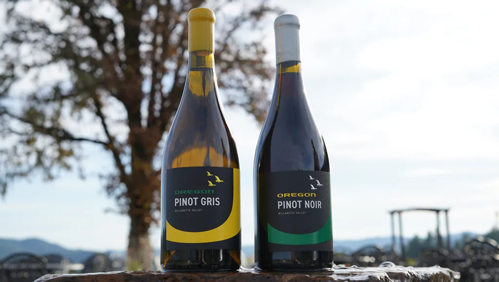 A UO-branded pinot gris and a pinot noir wine bottle stand next to each other against a blue sky background.