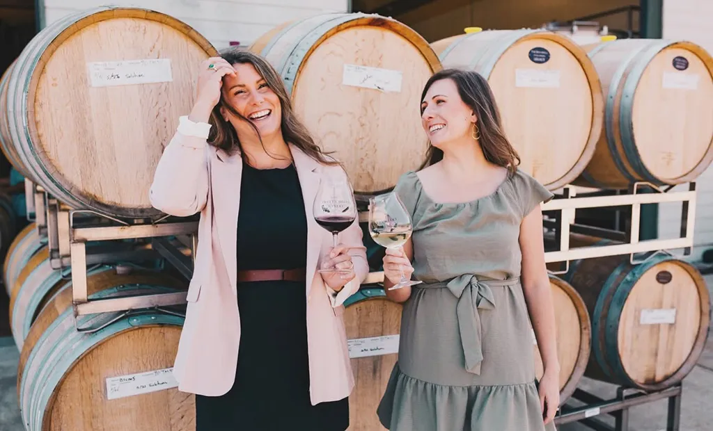 Two women holding wine glasses laugh in front of wine barrels.