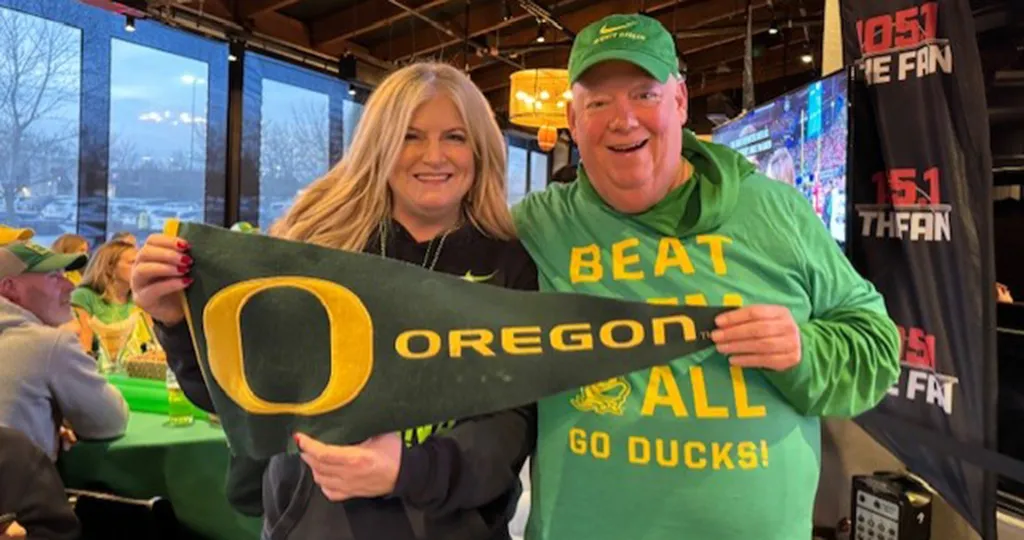 A man and woman in green Oregon gear hold up a University of Oregon pennant.