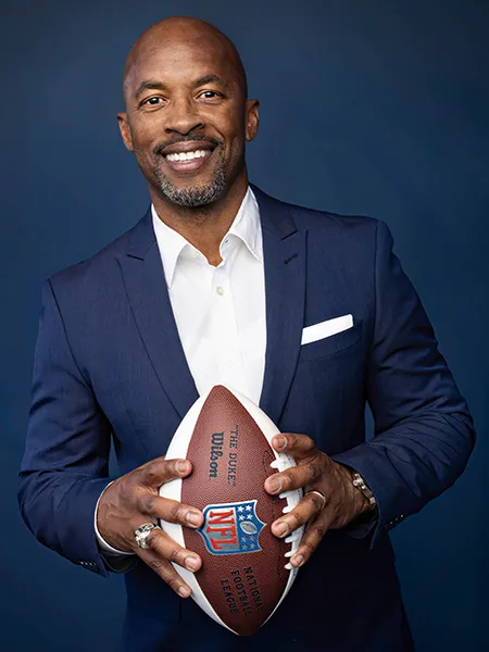 JJ Birden, a Black man in a dark blue suit holds a football with the NFL logo on the front.