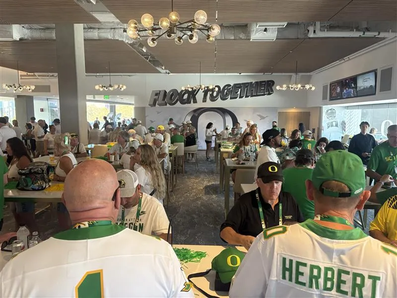 A packed room of Oregon Ducks fans in fan gear. The far wall reads in a silver graphic "Flock Together."