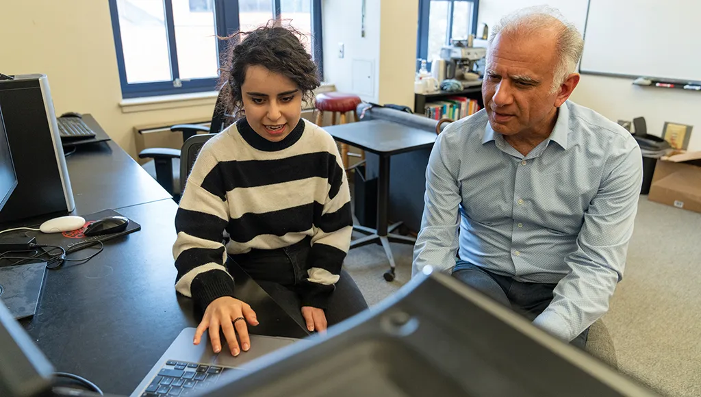 A south Asian woman sits next to an older man in a button up shirt as they both look at a computer screen.