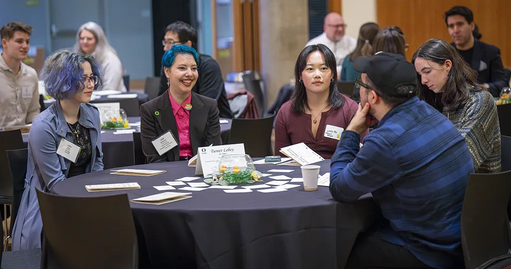 Four UO students smile while sitting around a table with a black tablecloth, facing a man in a blue flannel shirt. There are tables in the background with people talking around them as well.