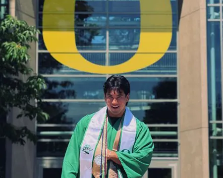 Logan Adams, wearing green graduation robes and two stoles, stands below a large yellow O sticker on the outside of Lillis hall.