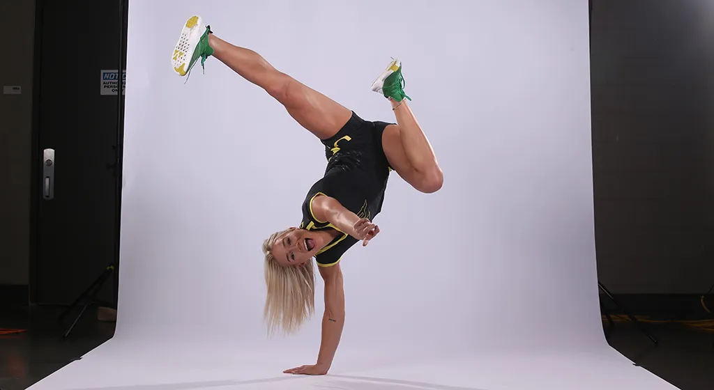 Calyx Hampton stands on one hand while pointing at the camera while smiling. One leg is extended out above her head and the other is bent toward her body.